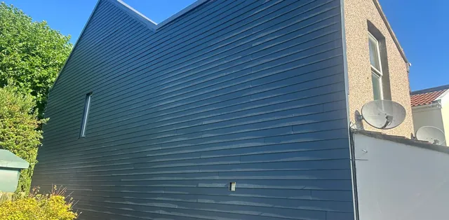 The gable end wall of a house covered by Pro Roofing in new dark grey cladding boards, in bright sunshine
